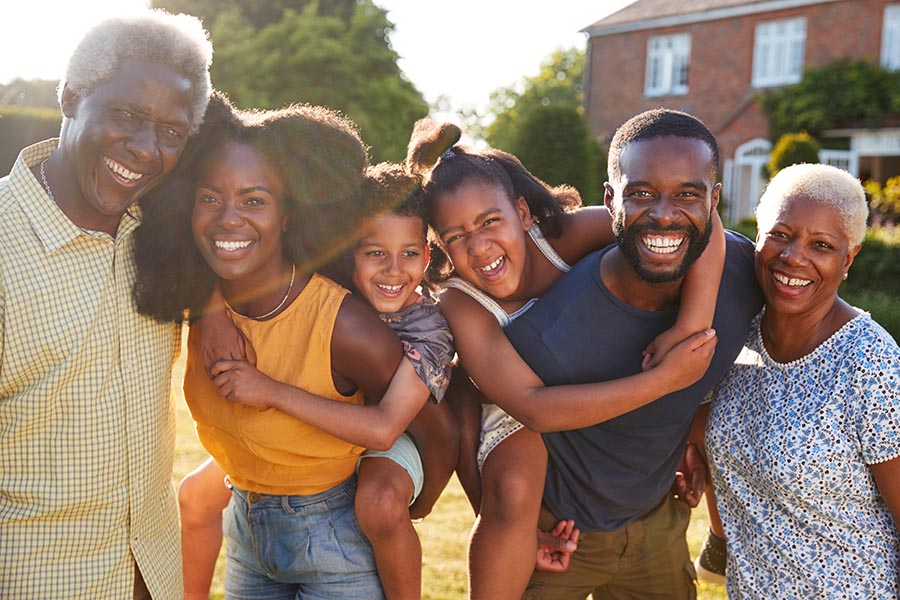 Personal Insurance - Multi-Generational Family Pose Outside Their Home, Children Riding on Their Parents' Backs, Everyone Smiling